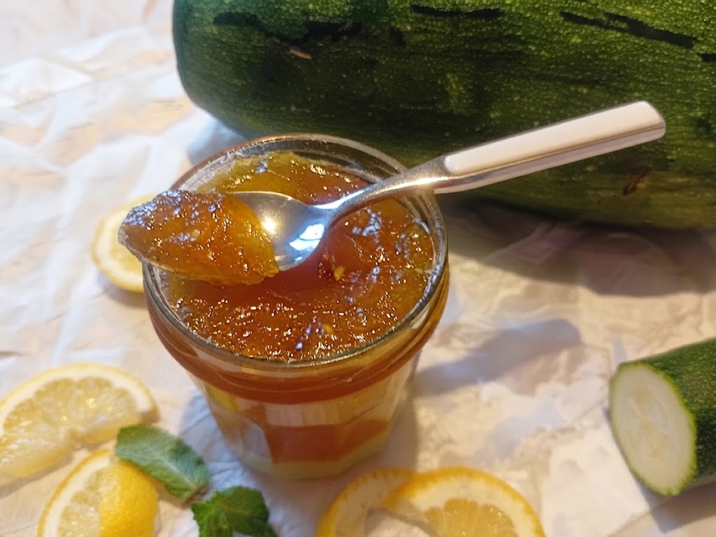 Un pot en verre rempli de confiture de courgettes au citron, d'une belle couleur ambrée, avec une cuillère à café posée dessus contenant une petite portion de la confiture. Autour du pot, sur une surface blanche froissée, sont disposés un économe, des rondelles de courgette, une demi-courgette entière en arrière-plan et des tranches de citron.