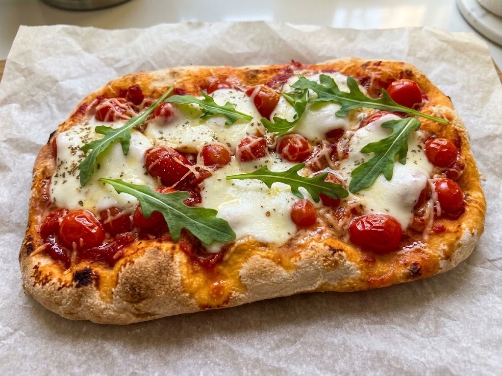 Une pinsa au jambon cru rectangulaire fraîchement cuite, garnie de mozzarella fondante, de tomates cerises rôties et de feuilles de roquette fraîche, reposant sur du papier sulfurisé froissé.