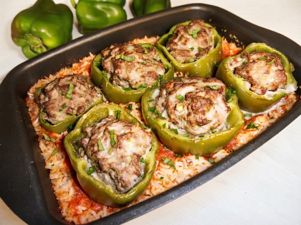 Plat de cuisson rectangulaire contenant six poivrons verts farcis de viande hachée gratinée, posés sur un lit de riz à la tomate au Cake Factory et garnis de basilic frais.